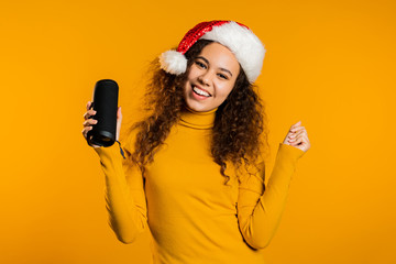 Young cute girl smiling and dancing with wireless portable speaker on yellow studio background....
