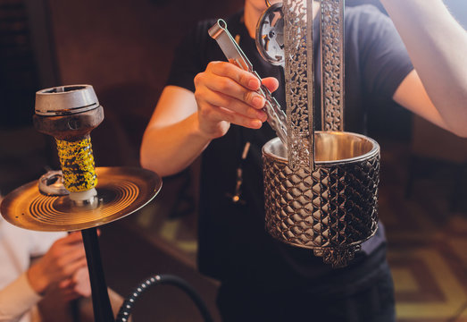 Hookah bowl with coal for smoking closeup. Hands holding forceps for shisha hookah and adjusts hot coals in metal bowl