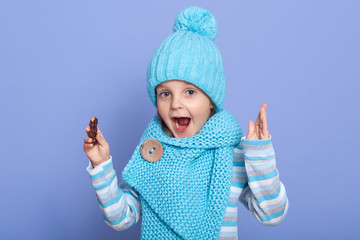 Close up portrait of little girl wearing winter warm turquoise cap and scarf posing over purple background, female child with opened mouth, looks astonished, being shocked, has pleasant look.