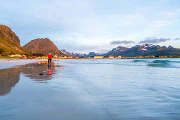 Fototapeta premium Man in red jacket stands on Ramberg beach in Lofoten island in Norway during sunset. Houses and mountains in the background. Travelocity and traveling concept.
