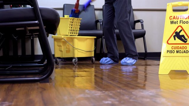 Woman janitor using a mop bucket and mop to clean the floor of hospital waiting room with medial protective clothing on