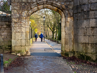 View through a stone arch to a couple strolling on the tree lined riverside Dame Judi Dench Walk in...