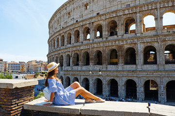 Travel woman in romantic dress and hat sitting and looking on Coliseum, Rome, Italy. Beautiful...