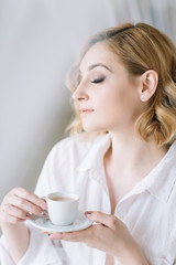 Beautiful young blonde woman in a light shirt in a bright room enjoys a morning cup of coffee before going to work.Girl in the bathroom in candid clothes.