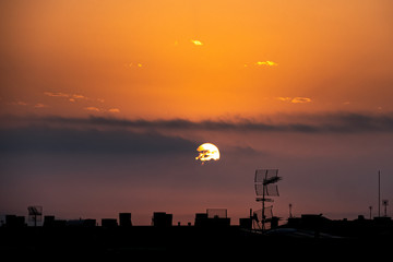 Fototapeta premium Sun appears from behind the clouds, above the city, roof top view. Summer sunrise or sunset. Orange sky color tones. Old town of Badalona, Spain