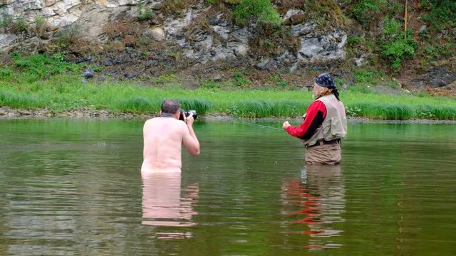 Fly fishing. A photographer with a naked torso waist-deep in water photographs a fisherman. in red clothes. Sports and entertainment on a mountain river.