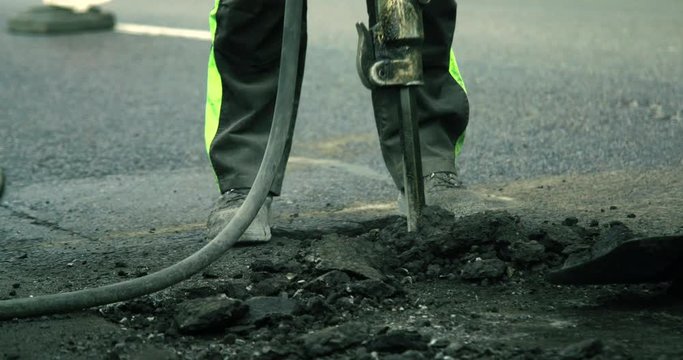 A construction worker is breaking the road with a jack-hammer while another worker is shoveling it away