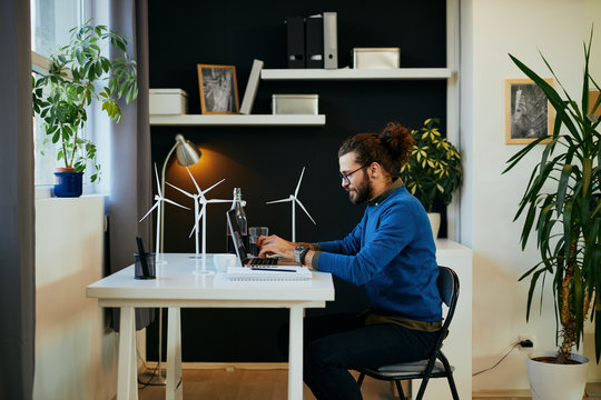 Young attractive bearded employee sitting in his modern office and using laptop for writing report about sustainability.