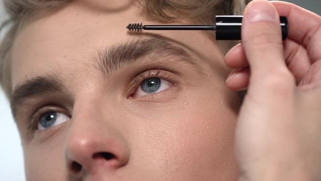 Macro of upper face of young attractive man with blue eyes and eyebrow brush coloring and brushing his eyebrow