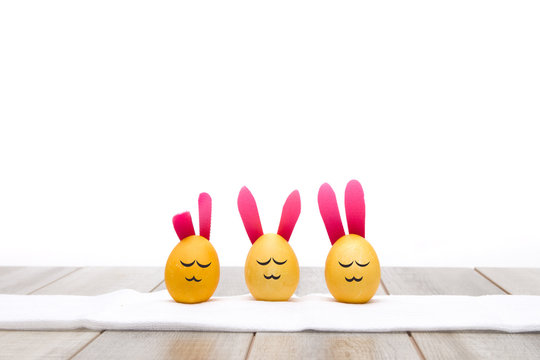 three yellow eggs with funny faces stay on a white towel with bunny ears on a white background, wooden table, copy space, easter card, place for your text