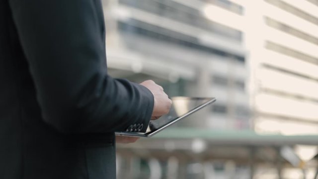 Close Up A Caucasian Office Worker In A Black Suit Using His Tablet Scrolling, Texting While Standing Outside On The Street Near A Big Office Building Urban.