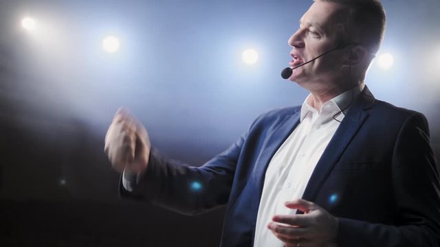 Showman. Middle Age Male entertainer, presenter or actor on stage. Arms to sides, smoke on background of spotlight. Front view portrait of a male public speaker speaking at the microphone, pointing
