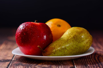 Naklejka na ściany i meble Fresh organic fruit red apple, orange and pear in white plate on rustic wooden table. glossy fruit with black background. Horizontal
