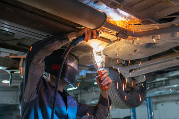 Car mechanic in welding mask welds car muffler