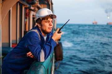 Marine Deck Officer or Chief mate on deck of offshore vessel or ship , wearing PPE personal protective equipment - helmet, coverall. He holds VHF walkie-talkie radio in hands.