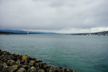 Tall white lighthouse on the shore of a bay with dark water against a cloudy sky in Geneva, Switzerland. Photography of an autumn sad landscape with cold sea water and a stone coast.