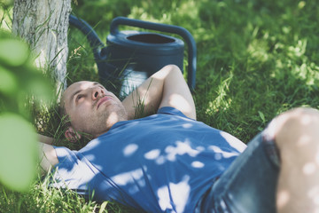 Man in blue t-shirt lying on grass with hands behind head in the garden with watering can nearby on greenery background on a sunny summer day. Free time and shying away from work concept. Toned