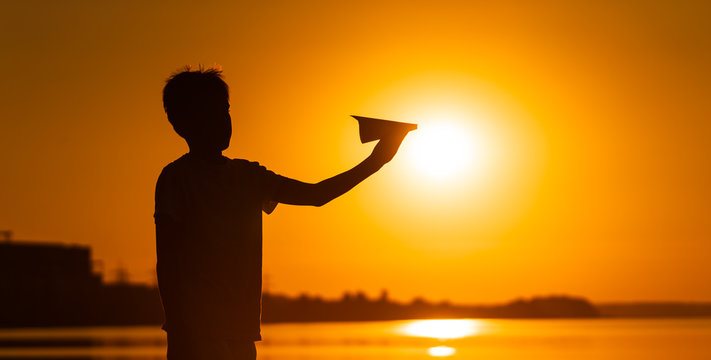 Black silhouette of a boy standing against orange summer sunset background. holding small paper plane in hands. Horizontal photo.