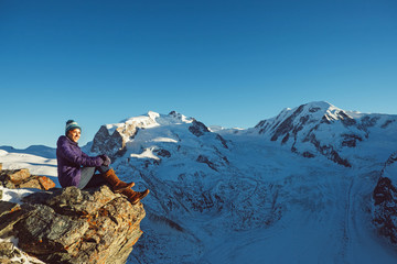 Fototapeta premium Happy traveler woman trekking in mountains, enjoy beautiful Matterhorn view. Explorer girl hiking on snowy hills, travel in Alps, Switzerland. Hiker sitting on rock cliff outdoors on nature.