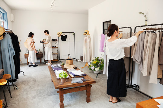 Three Japanese women standing in a small fashion boutique, looking at clothing on rails.