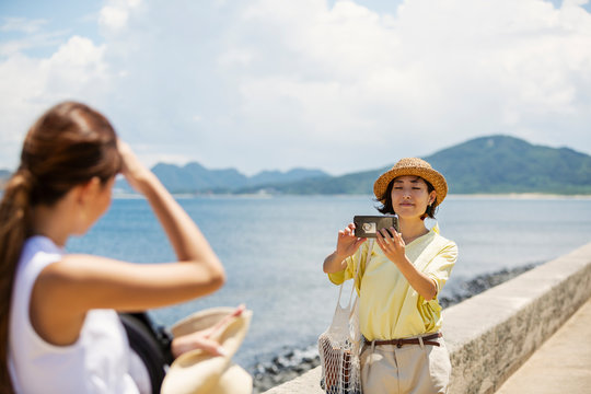 Two Japanese women standing by the ocean, taking picture with mobile phone.