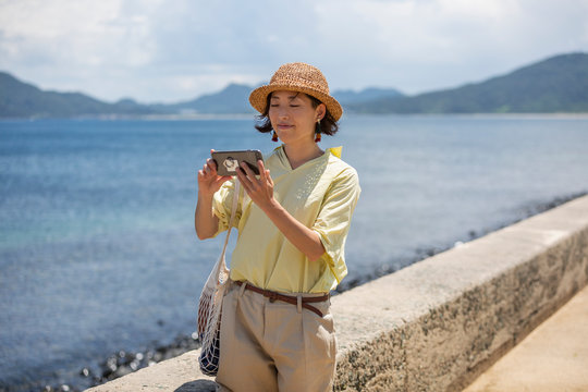 Japanese woman wearing hat standing by the ocean, taking picture with mobile phone.
