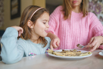 Cute Little girl sitting by the table while her mother is cutting the sweet pancake with cream daughter eating