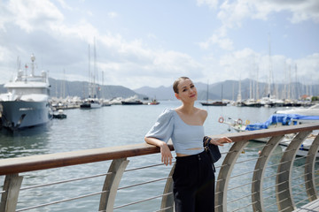 Fototapeta premium Young tourist woman on a bridge. Woman looks dreamy on the port with ships. Seascape with mountains. Summer vacation in Marmaris, Turkey. Copy space.