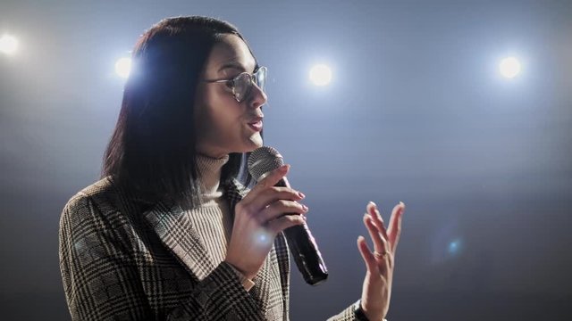 The Female Financial Coach Emotional Gesturing Talks From The Stage With Spectators At Forum. Too Many Anonymous Persons Workers And Students Seat In Large Auditorium And Watch Workshop Background