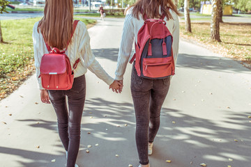 Two teenage girls walk in city on autumn day in summer, hold hands, look from back, casual clothes, jeans and a sweater, walk after school and college. Backpacks with textbooks and abstracts behind.