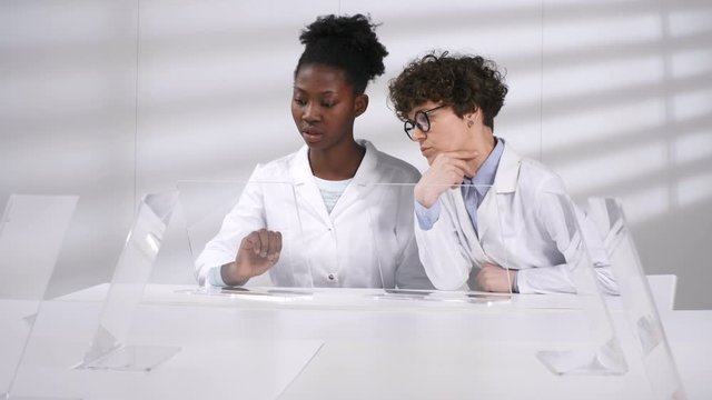 Waist-up shot of Caucasian and Afro-American female colleagues in white coats sitting together in lab and discussing computer simulation models on interactive AR screens. Template for AR, VR graphics.