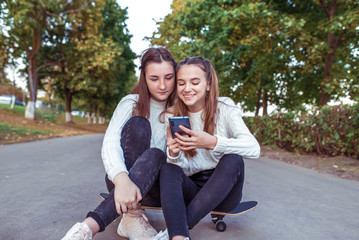 2 girls girlfriends 12-15 years old, happy sit smile, relax skateboard, city, rejoice and have fun. In hands of smartphone, watching videos online app social networks, casual clothes, jeans, sweater.