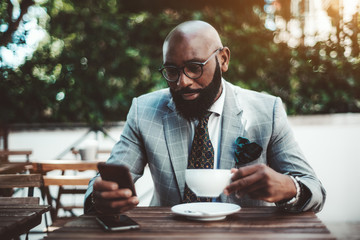 An elegant mature bald bearded African man in a fashionable costume with a necktie and spectacles is using one of his smartphones while sitting in a street cafe with a cup of a delicious tea