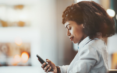 Side view of a dazzling mature African-American woman entrepreneur standing on the balcony indoors of an open-space area of an office and using her smartphone; a copy space place on the left - Powered by Adobe