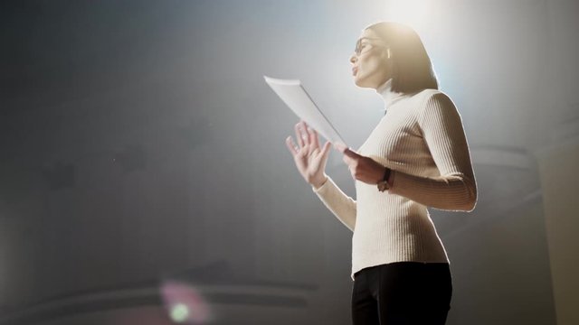 The Female Financial Coach Emotional Gesturing Talks From The Stage With Spectators At Forum. Too Many Anonymous Persons Workers And Students Seat In Large Auditorium And Watch Workshop Background