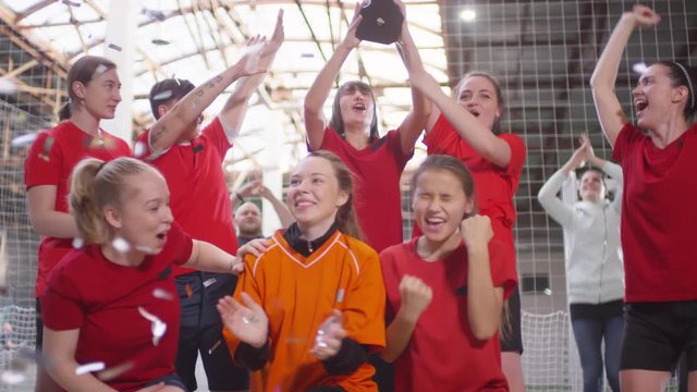 Team of happy female soccer players yelling in excitement, raising hands up and smiling while posing with cup in confetti shower after winning competition