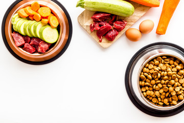 Pet feed ingredients. Raw meat and fresh vegetables near bowl with dry feed on white background top-down copy space