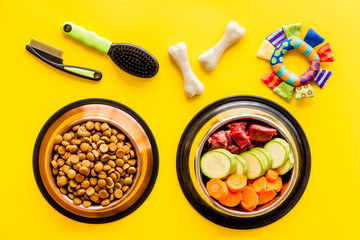 Dog feed - dry food and raw meat - near toy, brush and cheving bones on yellow background top view