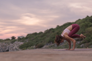 Young woman doing exercise yoga doing Crane (Crow) pose. Amazing yoga landscape in beautiful sky and enjoying sea view on rock floor, concept for exercising, health care.