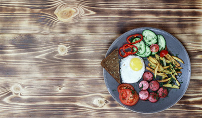 Breakfast - fried eggs, fried potatoes , tomatoes, cucumbers, pepers, sausage, toasts on plate over wooden background. View from above, copy space.