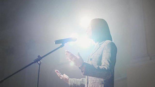 The Female Financial Coach Emotional Gesturing Talks From The Stage With Spectators At Forum. Too Many Anonymous Persons Workers And Students Seat In Large Auditorium And Watch Workshop Background