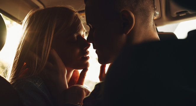 Close Up Portrait Of A Charming Young Couple Sitting Face To Face In The Car Trying To Kiss Against Sunset.