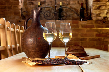 Two glasses of white wine, grape seeds, a clay jug and a few pieces of bread on a wooden table in a restaurant