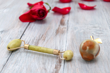 Jade roller for facial massage, jade apple and red rose petals on a gray wooden background....
