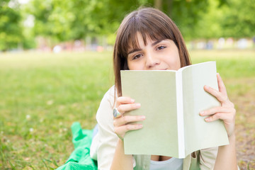Fototapeta premium Young woman reading book in park. Attractive young woman lying on lawn with book and looking at camera in park. Education concept