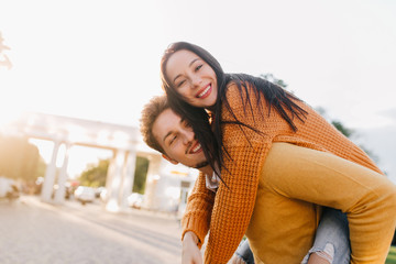 Glad man with beard in orange shirt carrying piggyback laughing girl. Excited young lady in knitted sweater riding on boyfriend's back in sunny morning.