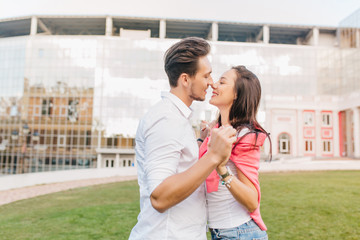 Stylish man with dark hair dancing with romantic girl on green lawn. Outdoor portrait of cute loving couple looking at each other with love and holding hands in front of modern building.