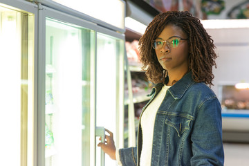 Young black female customer posing near fridge in grocery store. Buyer shopping in supermarket....