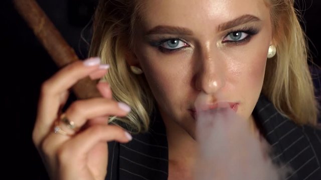 Close-up of the face of a seductive girl with bright make-up who smokes a cigar, she lets out a lot of smoke on a black background. Slow motion.