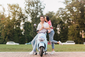 Fototapeta premium Glad girl in trendy jeans kissing boyfriend on scooter on blur nature background. Caucasian man in white clothes enjoying outdoor date with romantic young lady, standing beside him.